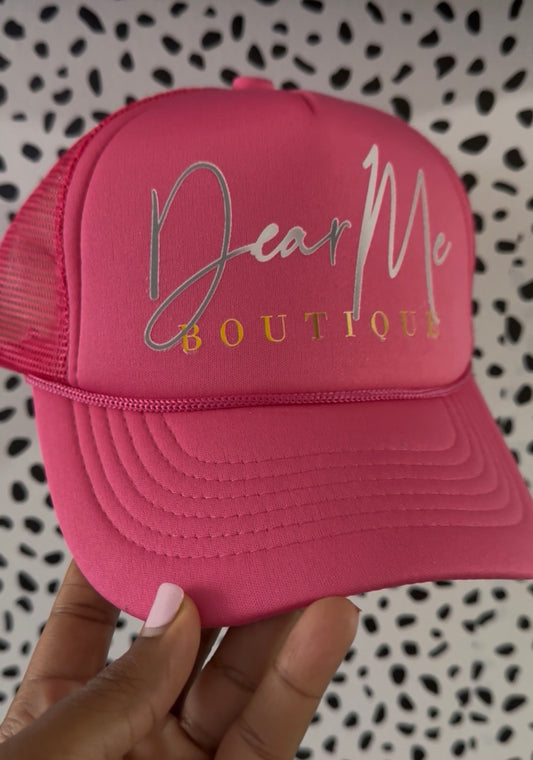 Pink cap with 'Dear Me Boutique' text held against a patterned background